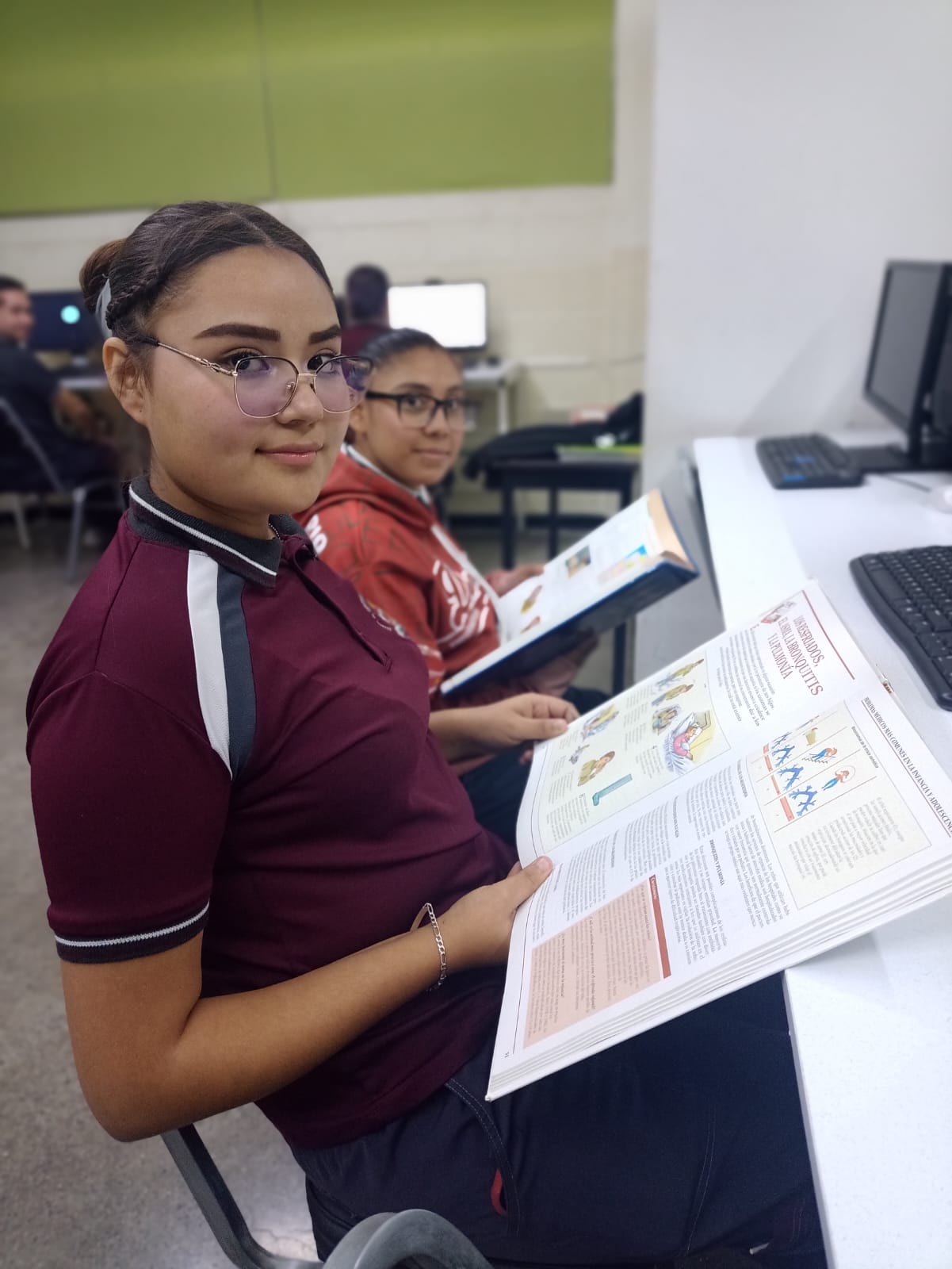 Estudiantes en uniforme guinda consultando material de estudio frente a las computadoras del aula.
