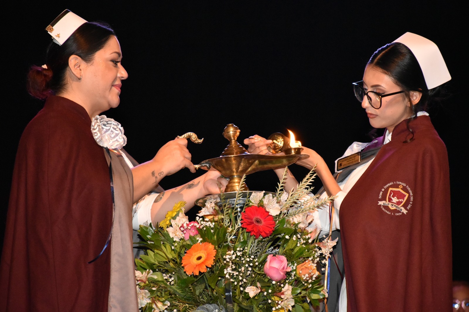 Ceremonia de la lámpara: una enfermera enciende la lámpara junto a una estudiante con cofia y capa.